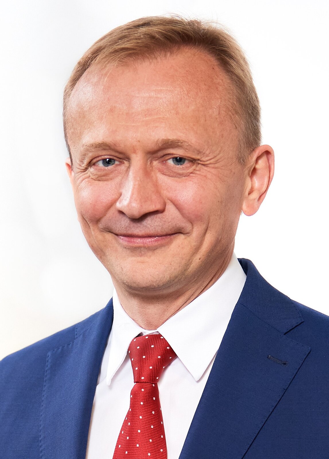 Fotografia przedstawiająca mężczyznę w średnim wieku. Postać ukazana jest od pasa w górę z twarzą skierowaną na wprost nas. Ma okrągłą twarz, rzadkie blond włosy i jest uśmiechnięty. Mężczyzna jest ubrany w białą koszulę i niebieską marynarkę. Na szyi ma zawiązany czerwony krawat w białe kropki. 