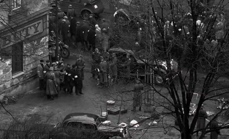 Zdjęcie przedstawia miejską ulicę. Stroją na niej tłumy cywilów w eskorcie niemieckich, uzbrojonych żołnierzy. Są także samochody i stojący przy nich niemieccy oficerowie.