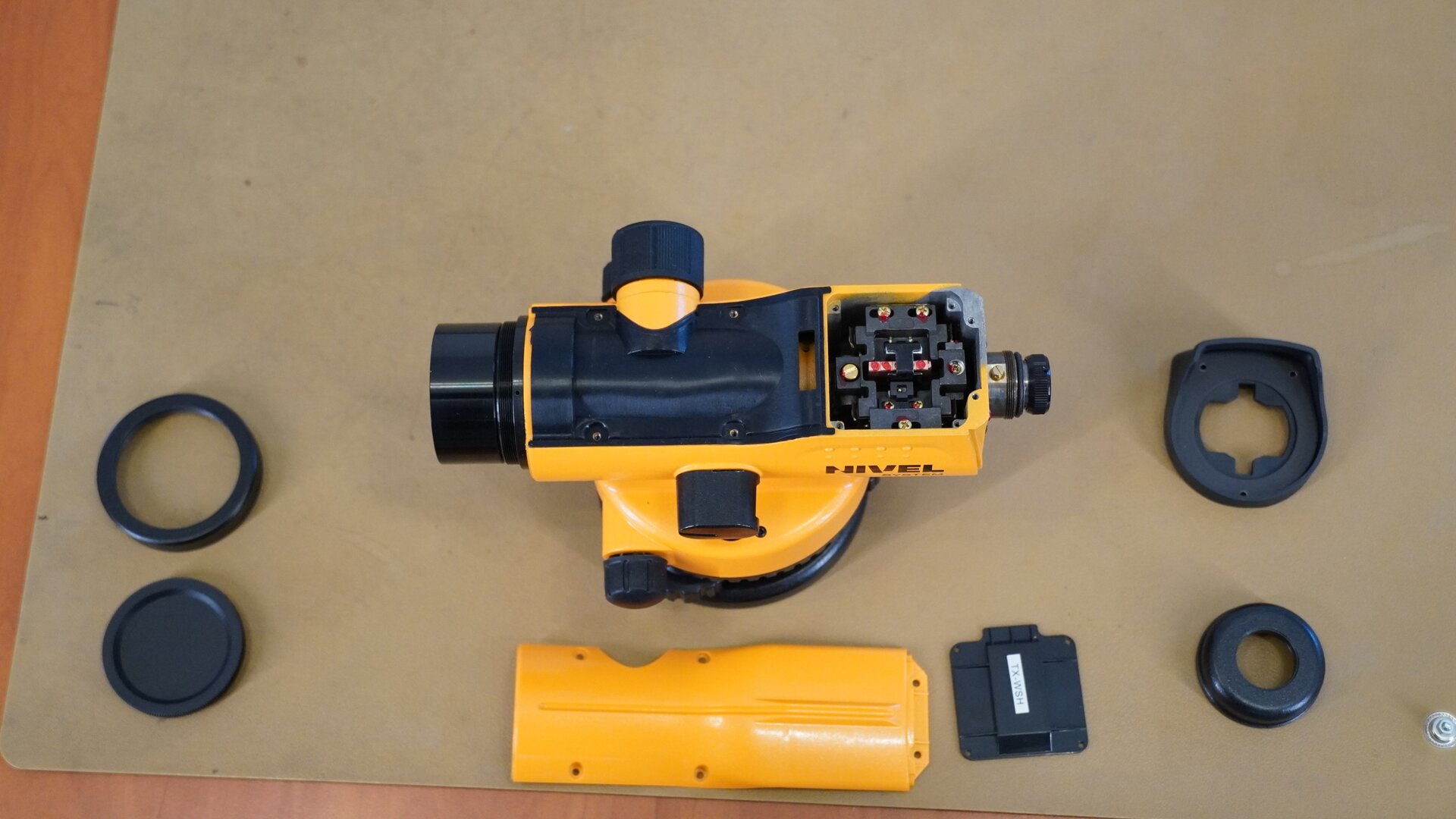 Zdjęcie przedstawia urządzenie widziane od góry. Zdemontowano z niego górną osłonę oraz obudowę okularu i osłonę kompensatora. Obok niwelatora umieszczono zdemontowane elementy: obudowę okularu złożoną z dwóch części oraz górną osłonę, obręcz obiektywu i jego przysłonę, a także osłonę kompensatora.