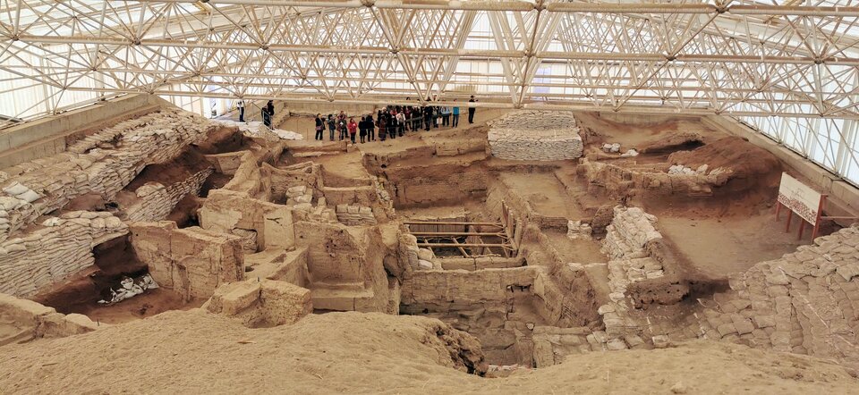 Fotografia przedstawia widok z góry na Çatalhöyük. Na pierwszym planie znajdują się ruiny, które pozostały po domach. Mają one grube mury i kształty nieregularnych prostopadłościanów. Za ruinami stoją ludzie i przyglądają się pozostałościom osady. Za nimi widać przeszkloną konstrukcję osłaniającą zabytek. 
