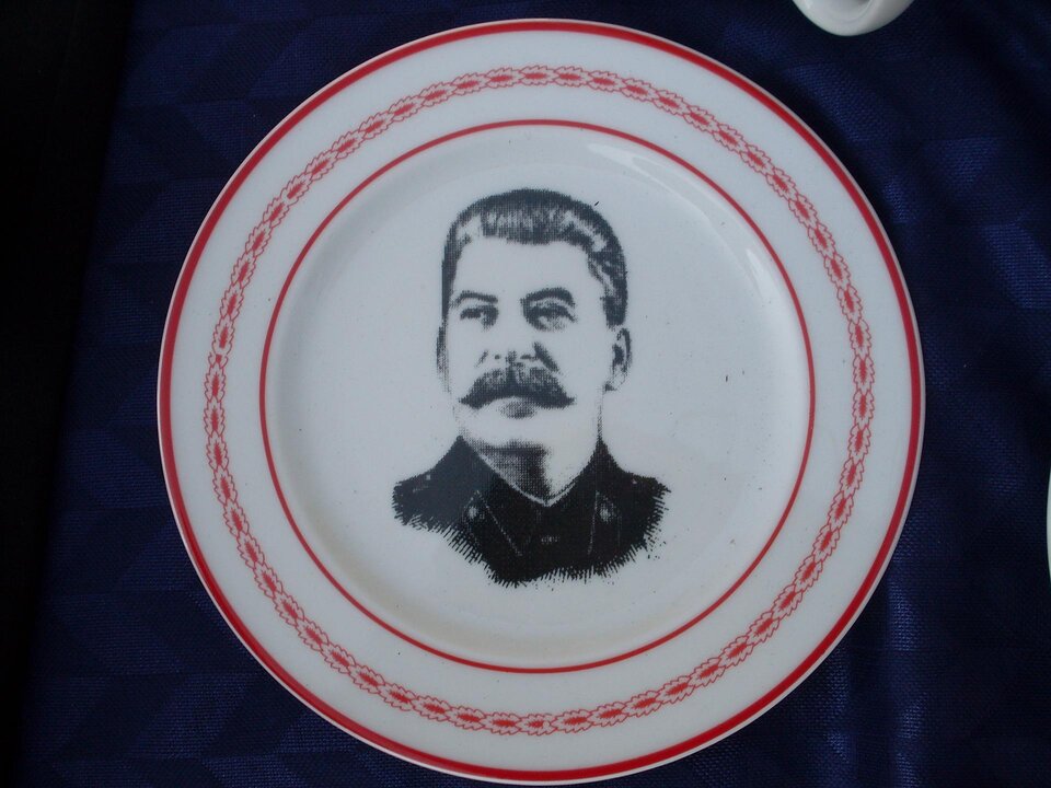 Zdjęcie przedstawia biały talerz z czarnym wizerunkiem Józefa Stalina. Widoczne jest jego popiersie. Ma gęste wąsy, zaczesane do tyłu włosy oraz ubrany jest w mundur. Talerz posiada czerwone obramowanie z prostym wzorem.