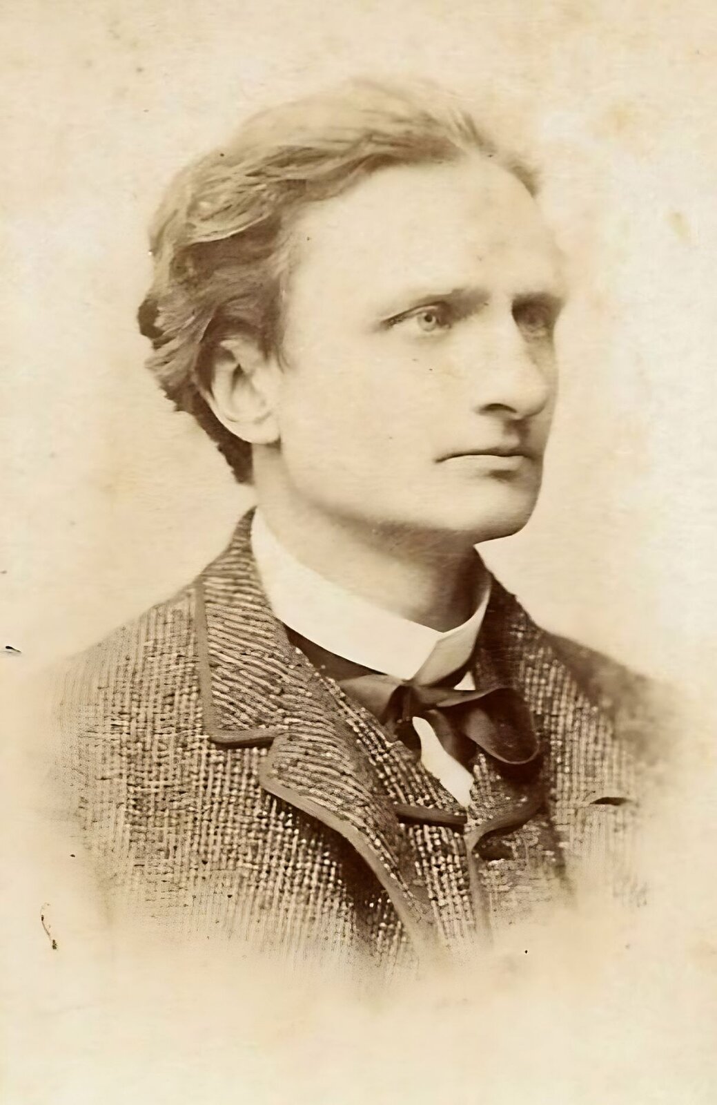 Zdjęcie przedstawia młodego mężczyznę z krótkimi włosami zaczesanymi do tyłu. Ma on długi, prosty nos i wąskie usta. Ubrany jest w melanżowy garnitur oraz kamizelkę. Pod spodem widoczny jest biały kołnierzyk koszuli oraz czarna wstążka przewiązana na szyi. Tło jest jednolite. Zdjęcie jest czarno‑białe.