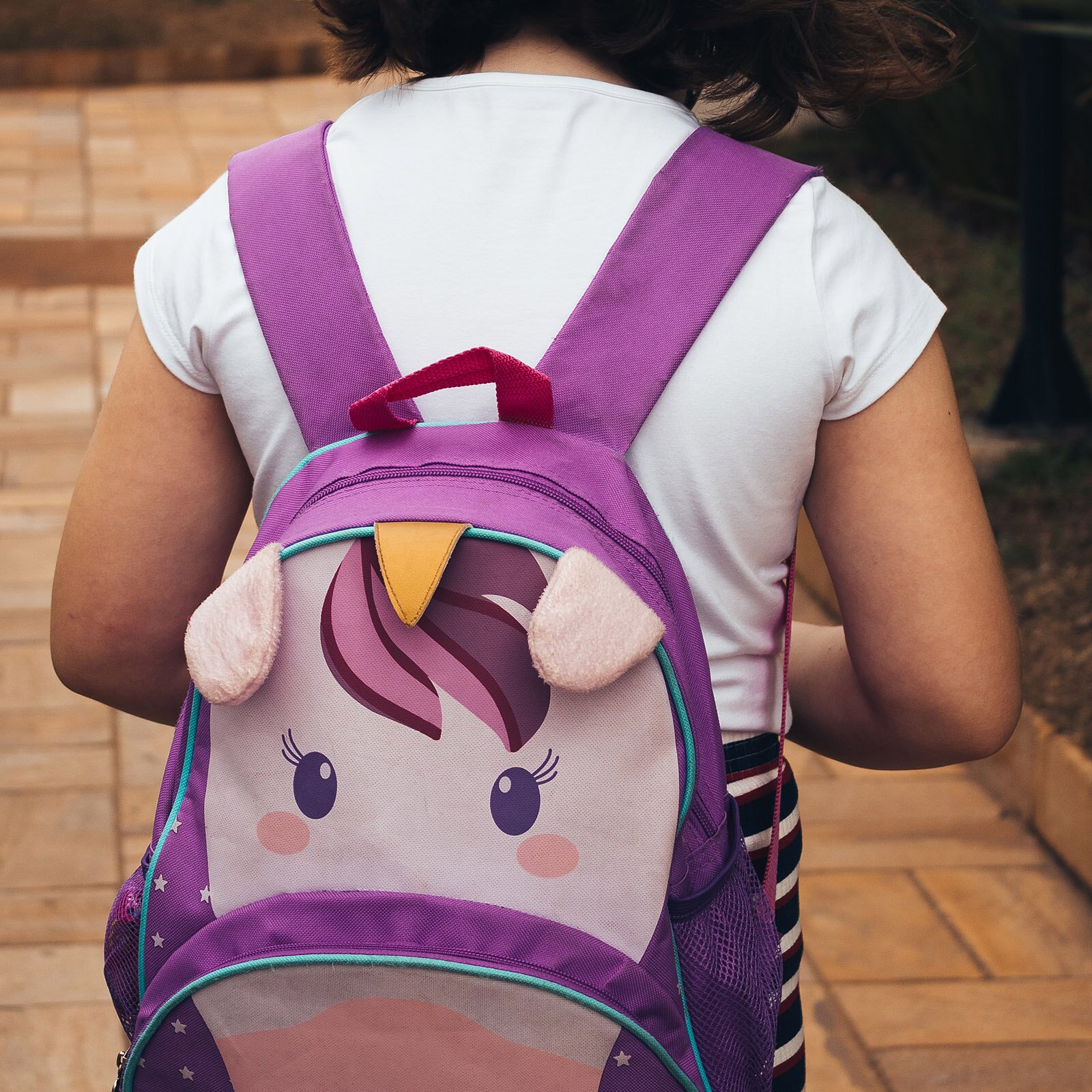 Zdjęcie przedstawia dziewczynkę od tyłu w wieku około 7 lat. Ma ona na sobie białą koszulkę i fioletowy plecak z podobizną głowy jednorożca. Plecak jest założony na oba ramiona dziewczynki, jego górna część zaczyna się pod łopatkami a dolna kończy prawie przy udach. 