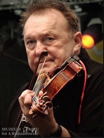 Mężczyzna grający na skrzypcach podczas występu; trzyma instrument pod brodą i prowadzi smyczek po strunach, skupiony, z widocznym oświetleniem scenicznym w tle.