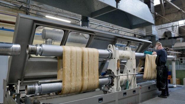 Zdjęcie przedstawia merceryzarkę wałową w hali zakładu włókienniczego. Maszyna jest otwarta. Na dwóch wałach zawieszone są tkaniny. Przy wale na dalszym planie pracownik w stroju ochronnym i rękawicach rozkłada tkaninę na wale. Za wałami ściana maszyny z szybami. Nad maszyną uniesione pokrywy. 