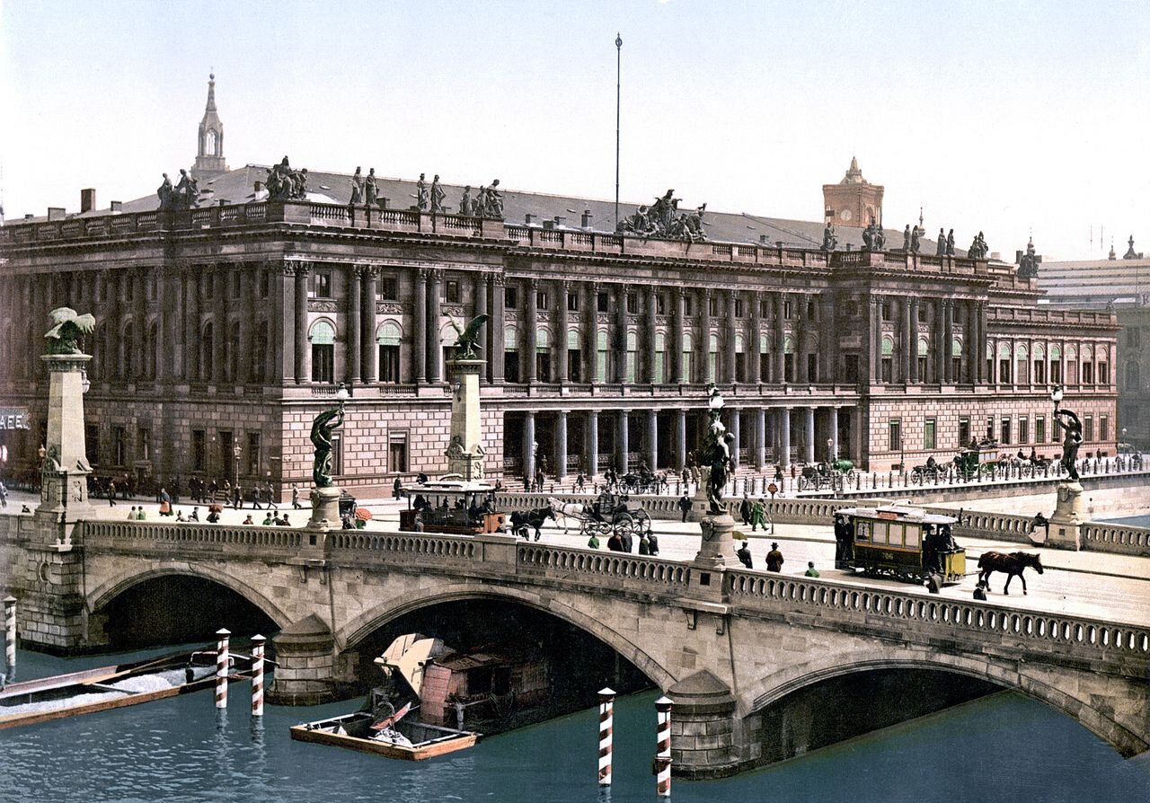 Zdjęcie przedstawia szeroki, kamienny, zdobiony rzeźbami most Fryderyka w Berlinie. Tuż za nim brzeg rzeki zdobi potężny gmach giełdy. Dwupoziomowy budynek , zwieńczony z każdej strony kolumnami. Na dachu tralki wraz z dekoracjami rzeźbiarskimi. Pomiędzy oknami sztukaterie. 