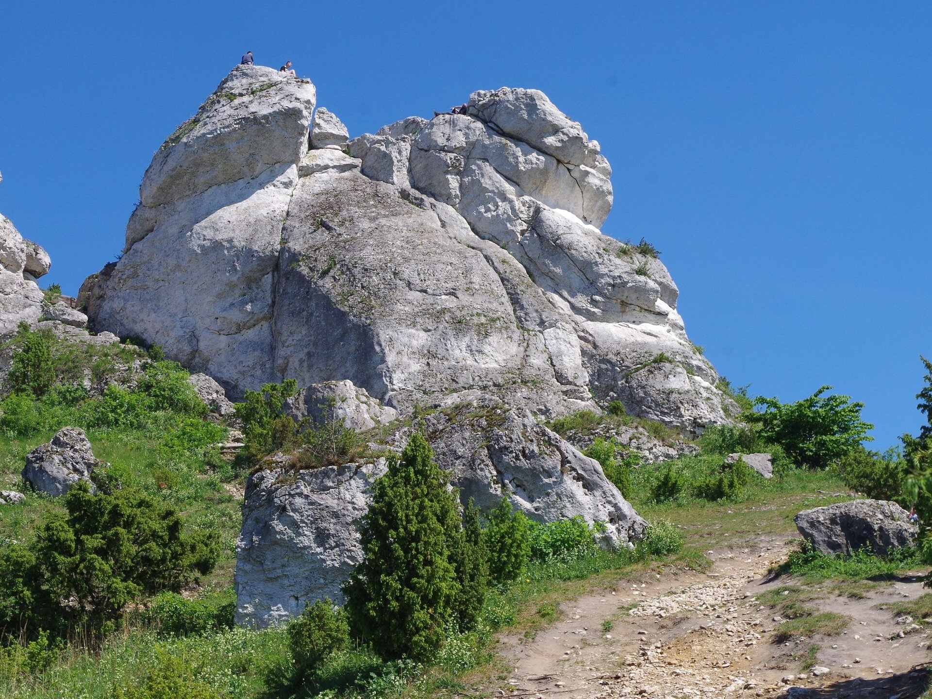 Fotografia przedstawia ostańca krasowego. To skalne wyniesienie w kolorze szarym i ciemnoszarym usytuowane na górzystym terenie, wyraźnie na wzniesieniu. W tle pogodne, niebieskie niebo z pojedynczymi chmurami.