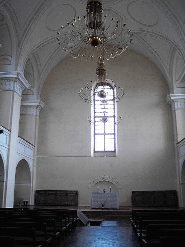 Ilustracja przedstawia wnętrze kościoła/katedry. Ściany są w kolorze bieli oraz beżu. Widoczne ozdobne żyrandole. Ołtarz jest skromny, na środku jeden prosty stół z dwoma świecami. Ławki dla wiernych w kolorze ciemnego brązu.