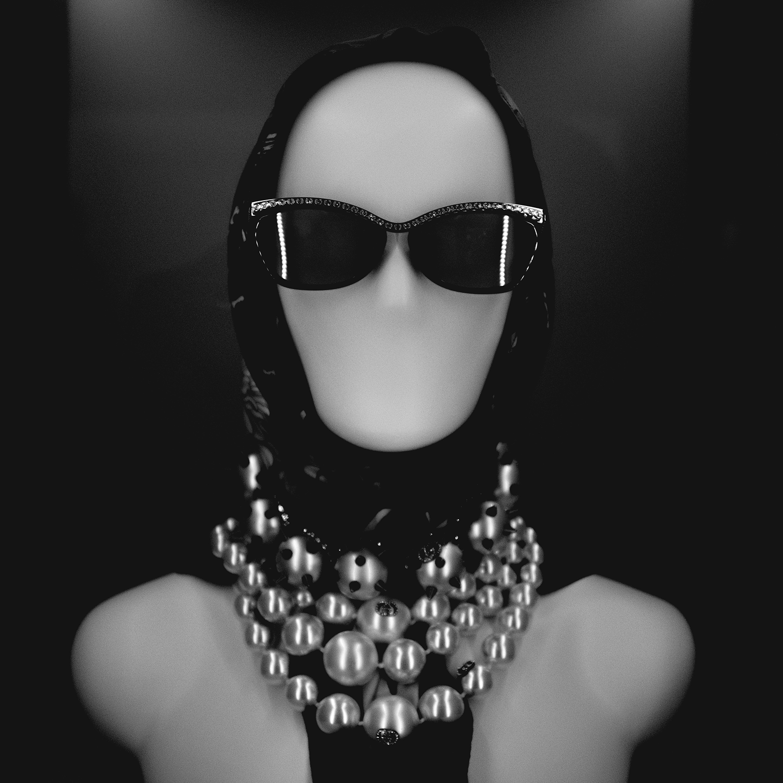 Zdjęcie przedstawia manekina mającego założone okulary przeciwsłoneczne, chustę na głowie oraz trzy rzędy pereł na szyi. 