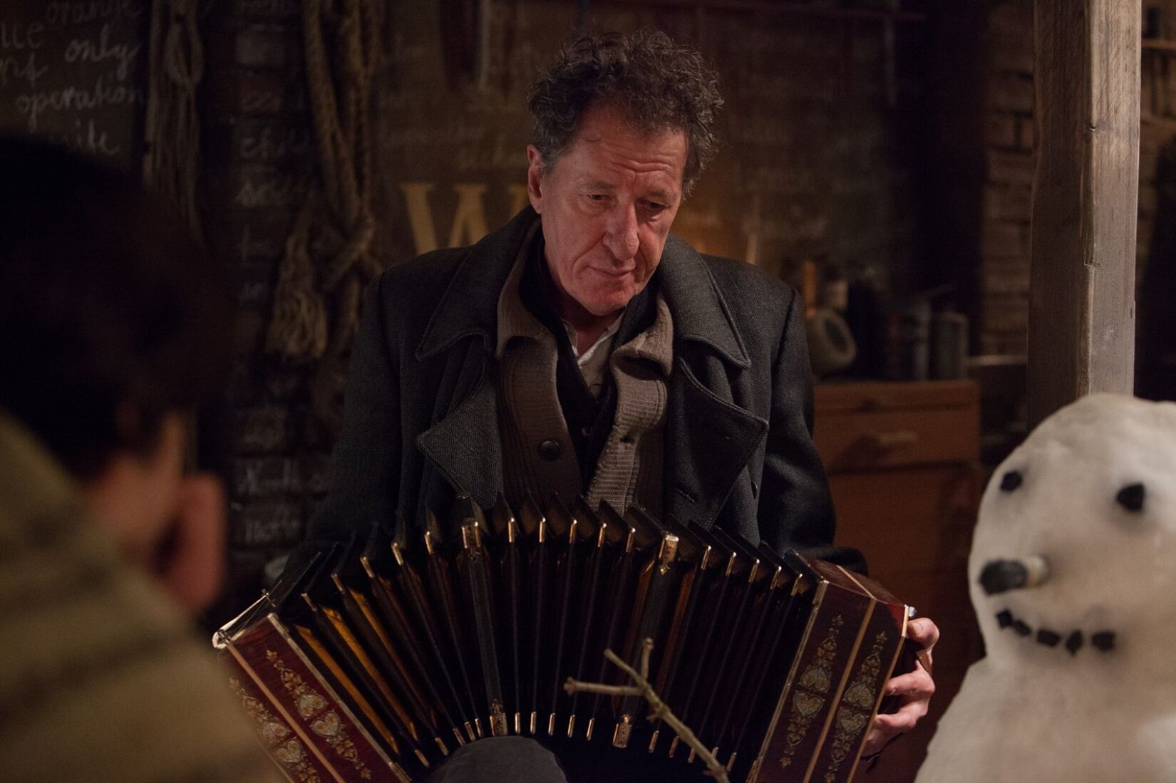 Zdjęcie przedstawia mężczyznę w średnim wieku grającego na akordeonie. Jest pokazany od pasa w górę. Ubrany w sweter i płaszcz. Ma krótkie kręcone włosy. Po jego prawej stronie stoi bałwan. W tle jest drewniana ściana.