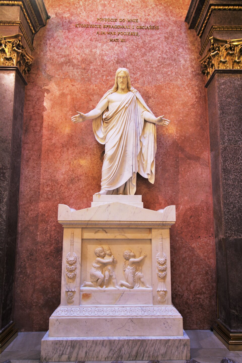 Zdjęcie przestawia rzeźbę prezentującą mężczyznę w sile wieku. Pochyla głowę. Postać ma owalną twarz, którą ukazuje w całości. Mężczyzna ma gładkie czoło, zamknięte oczy, prosty nos wąskie usta i zarośnięty podbródek. Włosy mężczyzny są długie, lekko falujące. Opadają na ramiona, są równo zaczesane na dwie stronu. Ma wyciągnięte na boki ręce z otwartymi dłońmi. Przez lewe ramię ma przewieszoną obszerną, pomarszczoną tunikę.