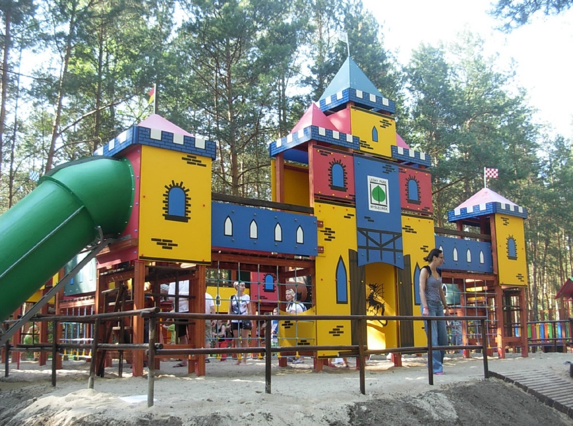 Ilustracja przedstawia plac zabaw w Leśnym Parku Kultury i Wypoczynku „Myślęcinek” w Bydgoszczy. Na zdjęciu ukazana jest drewniana, stylizowana na zamek budowla dla dzieci z drabinkami i zieloną, plastikową zjeżdżalnią. Elementy konstrukcji zwieńczone są spiczastymi daszkami z chorągiewkami. Na niebiesko‑żółto‑czerwonych ściankach namalowano okienka. Na dalszym planie znajdują się pozostałe elementy placu zabaw i wysokie, zielone drzewa. Całość otoczona jest drewnianym płotkiem.