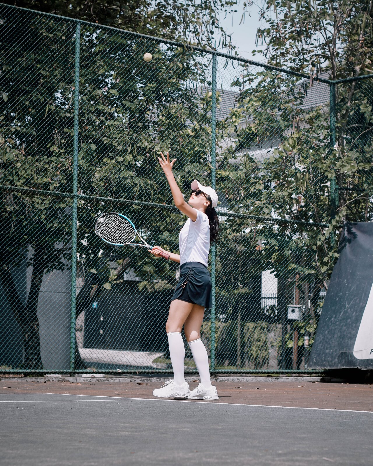 Zdjęcie przedstawia młodą kobietę na korcie tenisowym. Ma na sobie czapkę z daszkiem, koszulkę, luźne szorty oraz podkolanówki i sportowe buty. Dziewczyna w prawej ręce trzyma rakietę tenisową, a lewą podrzuca piłkę. 