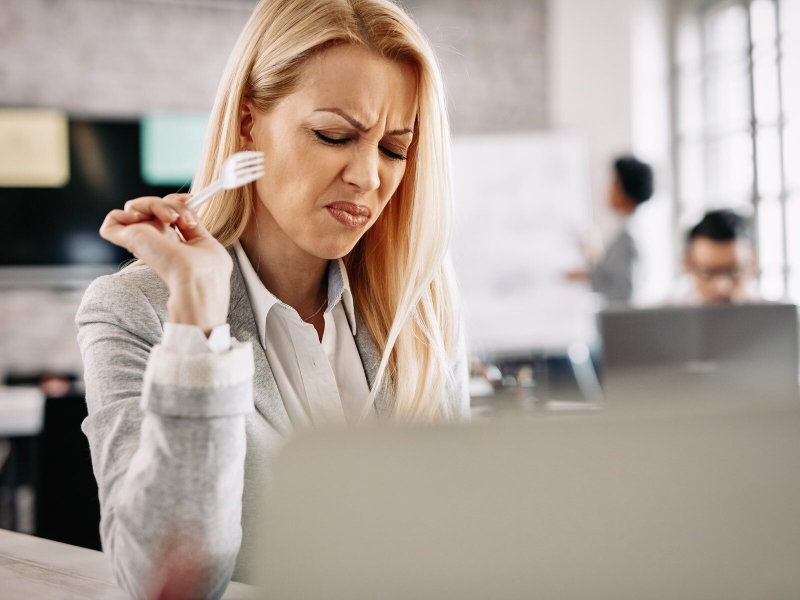 Zdjęcie przedstawia kobietę w średnim wieku, która siedzi w biurze i coś je. Kobieta ma długie, jasne włosy, jest ubrana w białą koszulę oraz szarą marynarkę. Zgiętą prawą rękę opiera łokciem o blat biurka, a w dłoni trzyma widelec. Ma mocno skwaszony wyraz twarzy. W tle widoczne jest rozmyte pomieszczenie oraz dwie postaci.