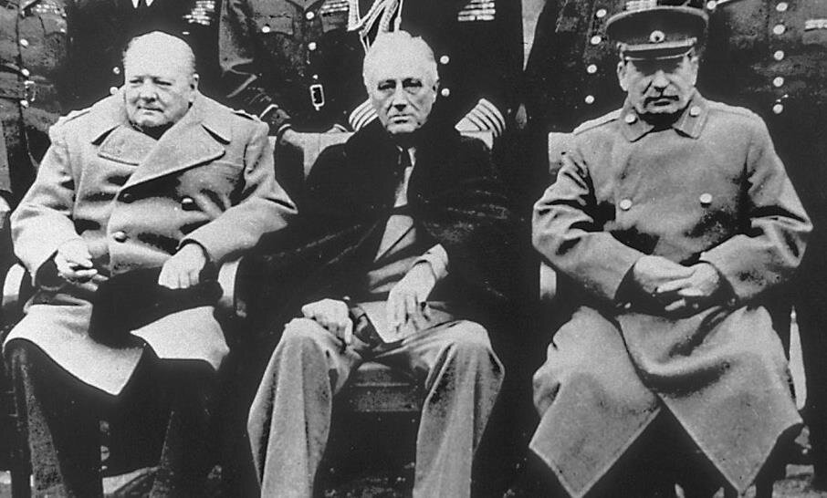 Czarno‑biała fotografia przedstawia trzech siedzących mężczyzn. Pierwsza postać od lewej jest ubrana w jasny płaszcz i ciemne spodnie. Na środku siedzi siwiejący mężczyzna, w jasnym garniturze i narzuconej na nim ciemnej pelerynie. Po prawej siedzi mężczyzna w czapce i mundurze Armii Czerwonej.