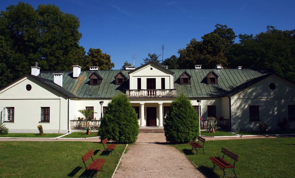 Na zdjęciu znajduje się biały dworek, zbudowany na planie prostokąta, z dwiema przybudówkami. Wykusz (element wystający z elewacji, nadwieszony powyżej parteru, podobnie jak balkon) wznosi się na dwóch  kolumnach z balkonem i marmurową balustradą. Na dachu znajdują się cztery lukarny (rodzaj nadbudówki skonstruowanej w dachu budynku) umiejscowione po dwóch stronach. W budynku dach jest spadzisty. Pokrywa go zielona dachówka. Są liczne,  białe, murowane kominy. Przed budynkiem znajduje się taras otoczony marmurową balustradą. Stoją na nim kwiaty. Do dworku prowadzi aleja, przy której stoją ławki. W tle znajdują się rozłożyste drzewa.