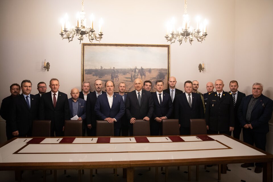 Zdjęcie przedstawia dużą salę, w której przy długim, prostokątnym stole stoi duża grupa mężczyzn w garniturach. Pozują do zdjęcia.