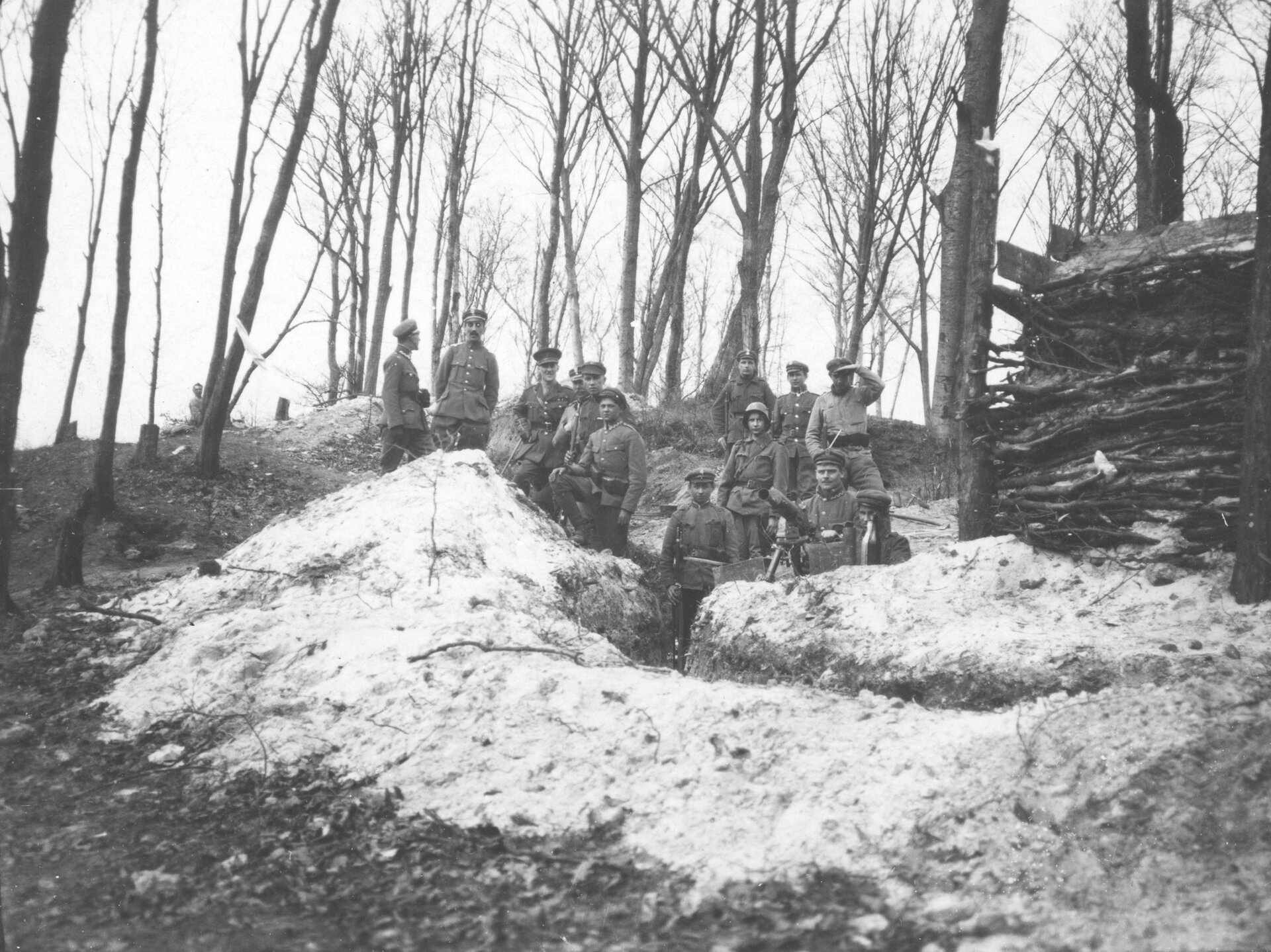 Zdjęcie przedstawia żołnierzy w okopie. Na wzniesieniu pokrytym śniegiem stoją żołnierze w mundurach i czapkach. Jeden z nich podtrzymuje karabin maszynowy. Z prawej strony jest stos ułożonych jedna na drugiej gałązek. Wokół są nagie drzewa.