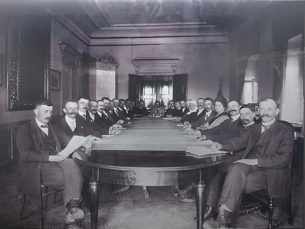 Fotografia przedstawia grupę ludzi, głównie mężczyzn siedzących przy długim stole, patrzących w obiektyw. Mają przed sobą rozłożone papiery. Wszyscy są elegancko ubrani, w garnitury, koszule, krawaty lub muchy. Wśród nich są dwie kobiety. Na ścianie po lewej stronie widać duży obraz ze zdobioną ramą. W szczycie stołu widać drewniane drzwi, po prawej stronie dwa okna.