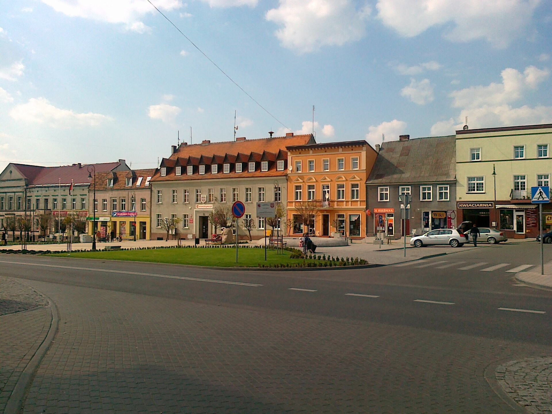 Kliknij, aby powiększyć. Zdjęcie przedstawia fragment miasta. Domy są jedno, dwu- i trzypiętrowe. Są różnokolorowe, a okna umieszczone są w nich symetrycznie i w równych odległościach. Przylegają do siebie, tworząc zwartą zabudowę. Część domów ma skośne dachy, a inne płaskie. Przed budynkami deptak wyłożony chodnikiem, a także trawa znajdująca się na jego rogu. Na deptaku są ławki, siedzą na nich ludzie. Przy trawie ulica, a przy niej parking, znajdujący się z prawej strony, na którym stoją samochody. Ulica i deptak znajdują się na pierwszym planie, natomiast zabudowa na drugim.