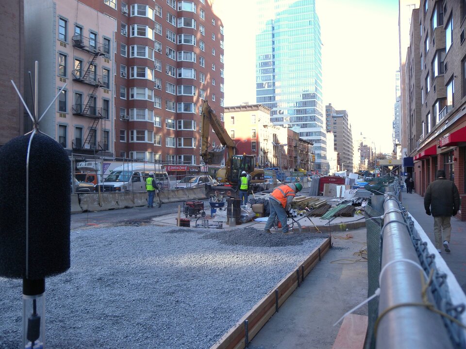 Zdjęcie przedstawia roboty drogowe w mieście. Wykonywane są pomiędzy wysokimi budynkami. Na pierwszym planie jest nawierzchnia wysypana drobnym, jasnym kamieniem. Pracuje przy niej kilku robotników.   