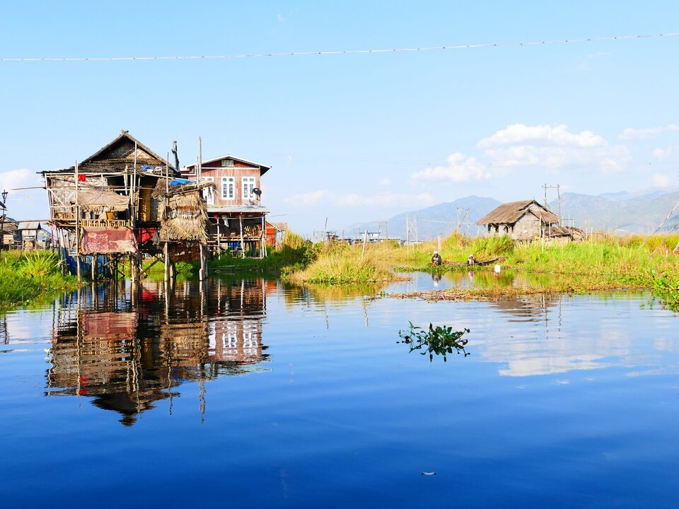 Zdjęcie przedstawia drewniane, ubogie domy stojące na palach. Pale są częściowo zanurzone w wodzie. Gdzieniegdzie teren porasta roślinność przypominająca trzcinę. W tle są góry. Niebo jest bezchmurne.  