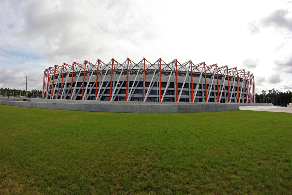Ilustracja przedstawia stadion Miejski w Białymstoku. Budynek na planie owalu, z biało‑czerwonymi elementami elewacji, wykonanymi z metalu.