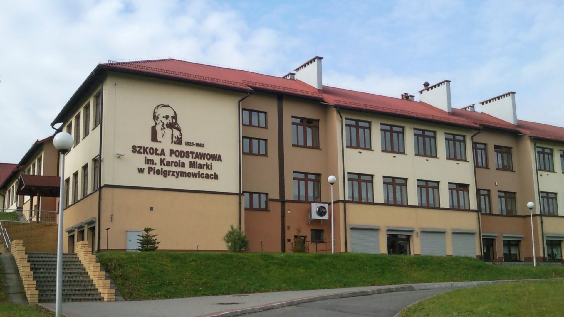 Fotografia przedstawiająca budynek szkoły podstawowej imienia Karola Miarki w Pielgrzymowicach. Jest to budynek trzykondygnacyjny, do którego wchodzi się po schodach.Na fasadzie budynku umieszczono portret patrona szkoły.