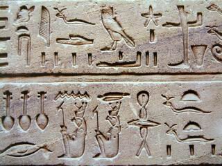 Fotografia przedstawia hieroglify egipskie wyżłobione na gładkiej powierzchni. Na dwóch oddzielonych od siebie linią płaszczyznach widnieją wyryte w skale skomplikowane symbole, między innymi symbol ślimaka, sowy, pióra, stopy oraz siedzącej postaci ludzkiej.