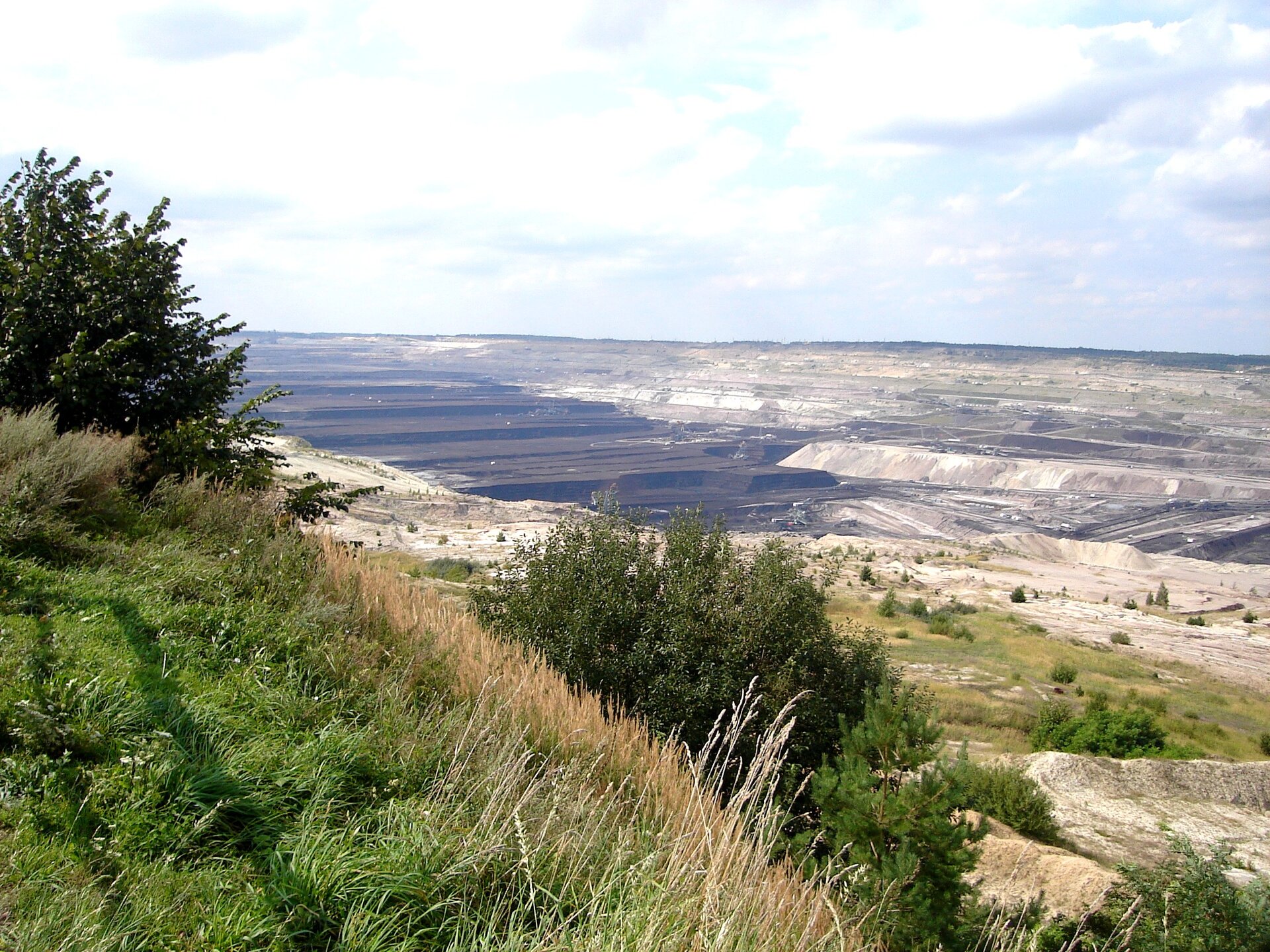 Zdjęcie przedstawia rozległą kopalnię odkrywkową. Teren kopalni jest położony w obniżeniu terenu. Ma liczne tarasy ciemnego koloru.  