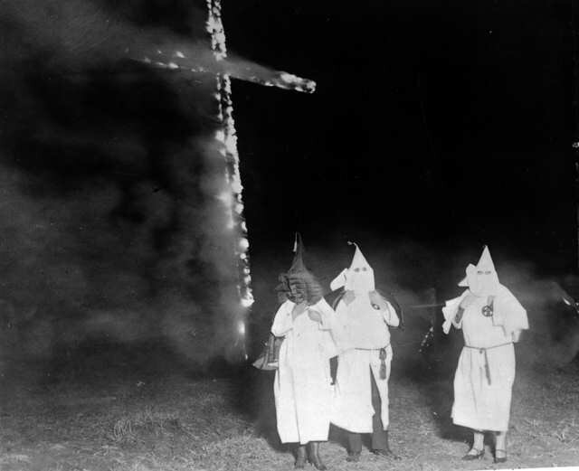Ilustracja przedstawia scenę z udziałem członków Ku Klux Klanu. W centrum widać płonący krzyż. U jego stóp trzy postaci odzianych w stroje Ku Klux Klanu- białych płachtach materiału do kostek i białych kapturach z wyciętymi dziurami na oczy. Kaptury skrywają całą głowę i są zakończone trójkątnym, stojącym szpicem. W stroje są ubrane dwie postacie żeńskie i męska, stojąca pośrodku pomiędzy kobietami. Można to zauważyć po obuwiu. Dwie postaci mają buty na obcasie, męska półbuty. Scena rozgrywana wieczorem. Zdjęcie czarno‑białe. Na członków klanu pada światło z reflektora. 