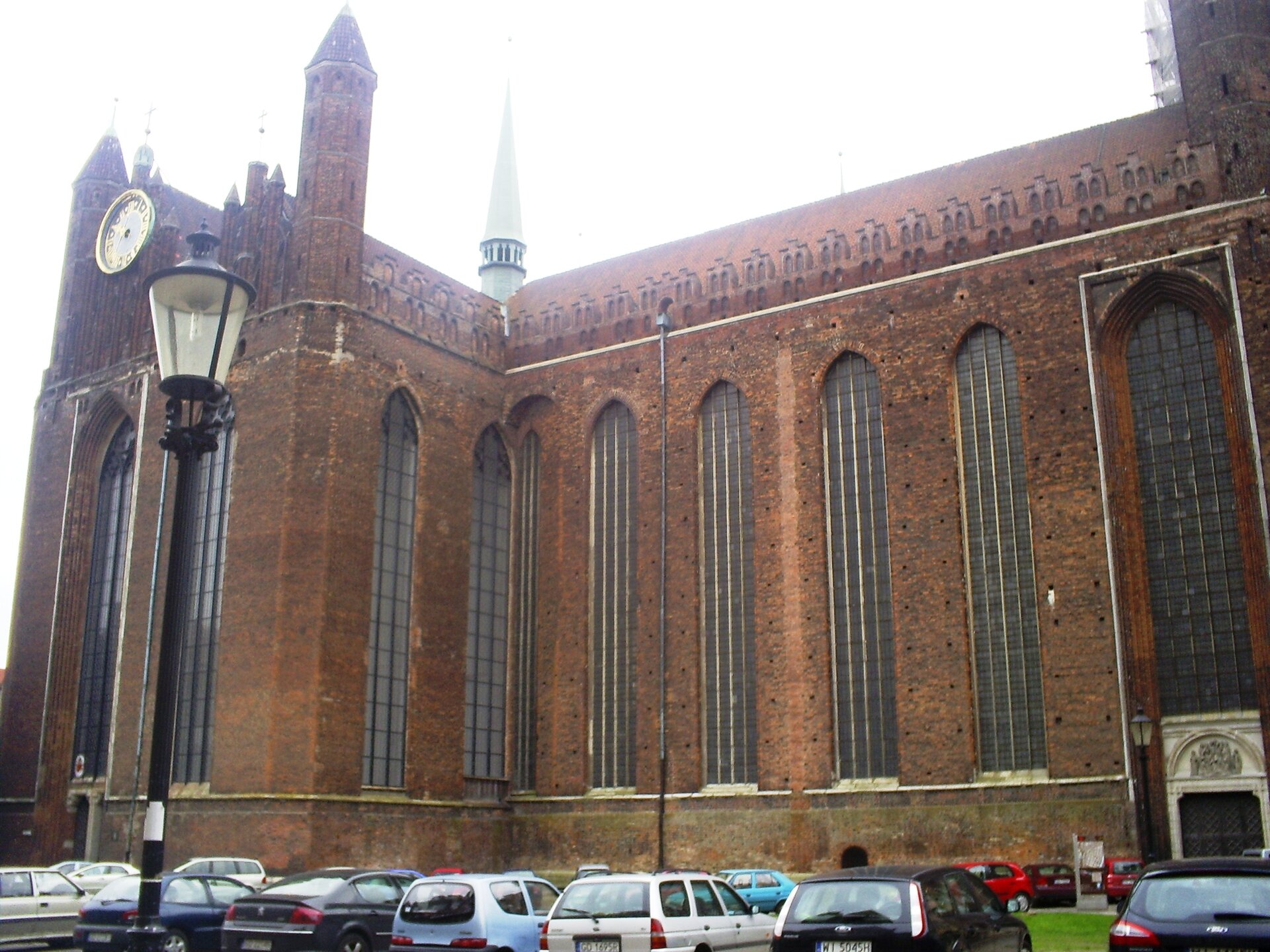 Zdjęcie przedstawia widok współczesny na ścianę zewnętrzną nawy bocznej Kościoła Mariackiego w Gdańsku. Budynek wykonany z cegły, portale drzwi wejściowych wykonane z kamienia. Na całej wysokości ściany widoczne są wbudowane wysokie, ostrołukowe okna witrażowe. Na szczycie dwuspadowego dachu widać gotycką stylizację wzdłuż całej długości obwodu dachu. Po lewej stronie wieżyczki ceglane flankują górną część zabudowy dachu, na którym umieszczono zegar tarczowy. Widoczna na szczycie dachu sygnaturka, uchwycona z perspektywy obiektywu w centrum fotografii. U dołu ilustracji parkujące na placu parkingowym pod kościołem samochody.