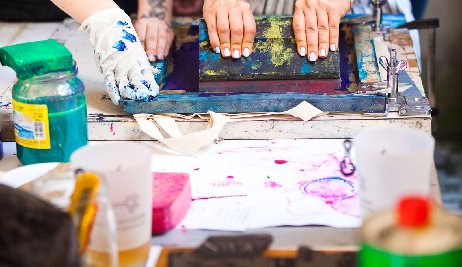 Zdjęcie przedstawia moment pokrywania matrycy farbą drukarską. Na stole leży matryca, a za nią 2 pary rąk, jedne, w gumowych rękawiczkach przytrzymują matrycę , druga osoba nanosi farbę za pomocą szerokiej szpachli. Obok stoi słoik z farbą, na którym leży gąbka. Przed matrycą leży używany, trochę przybrudzony farbą papier, z brzegu niewyraźnie widoczne pojemniki i butelki. 