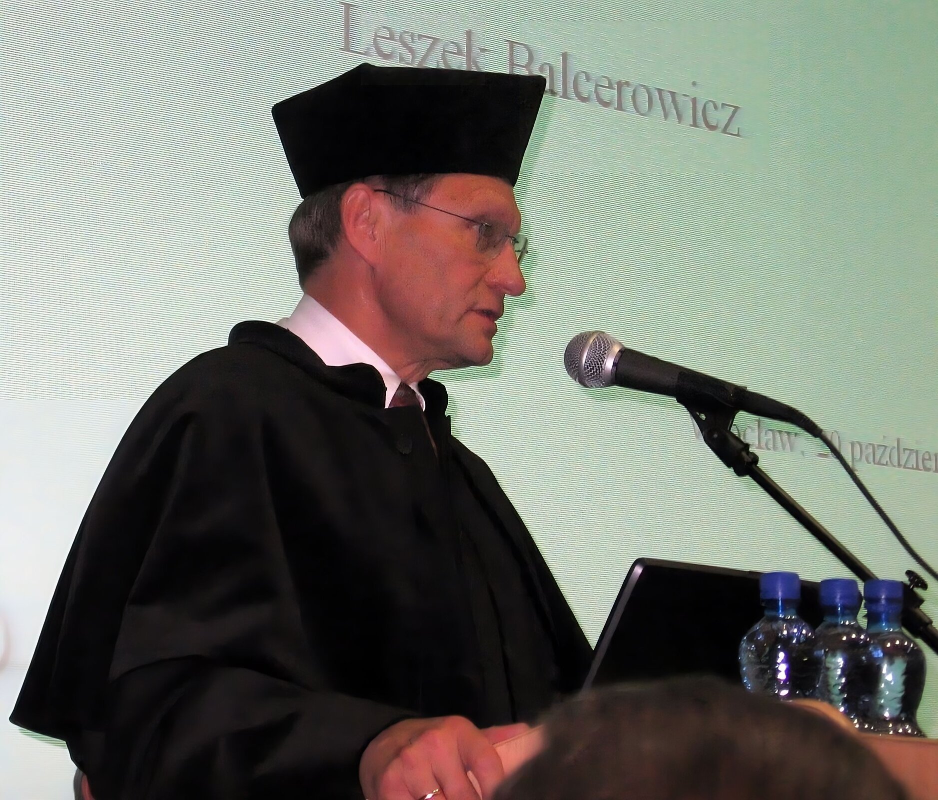 Mężczyzna w średnim wieku, ubrany w czarną czapkę i czarną szatę, w okularach mówi do mikrofonu. Z tyłu ekran, na którym wyświetlany jest napis: Leszek Balcerowicz, Wrocław, 20 paźdźiernik.