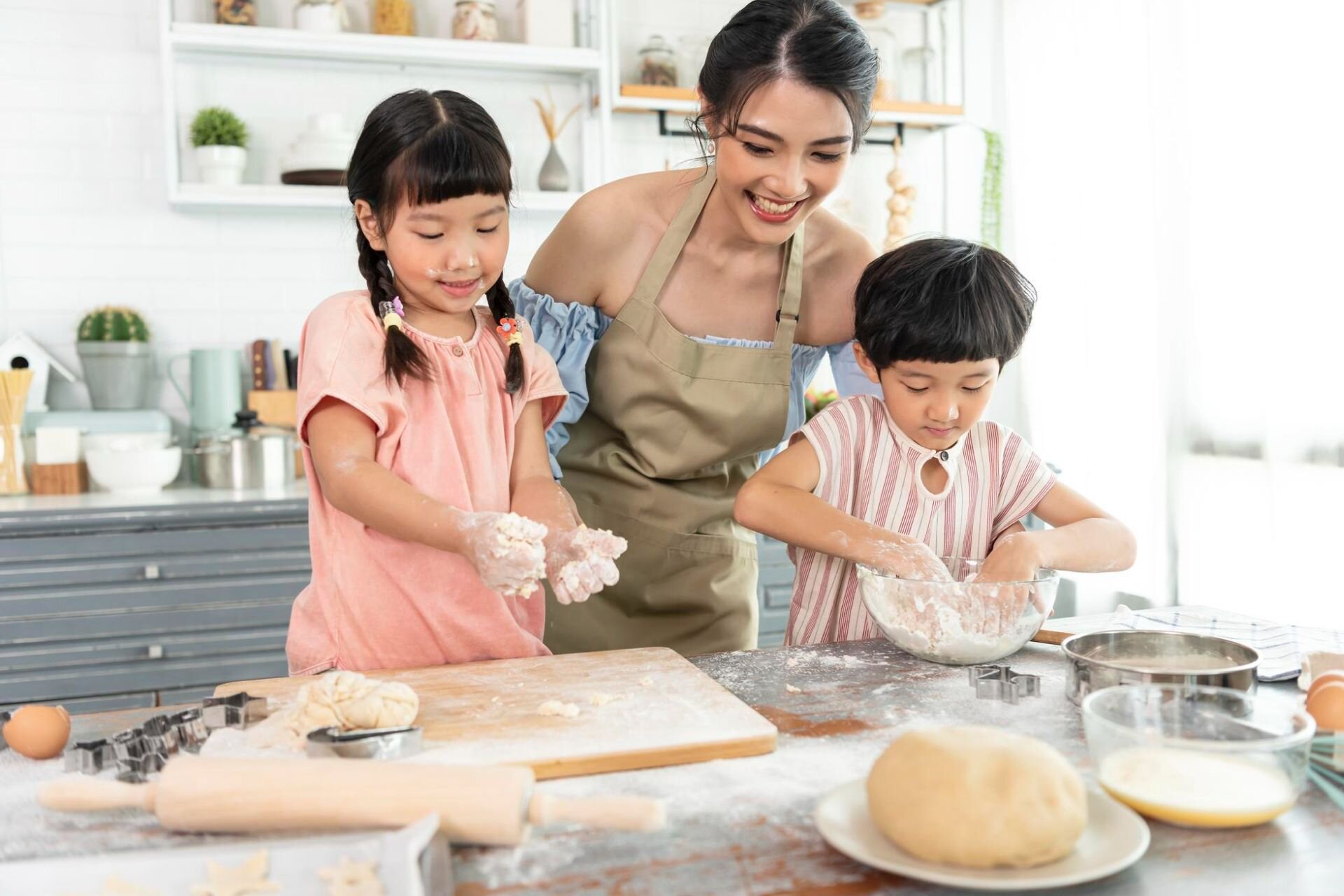 Ilustracja przedstawia matkę i dzieci piekących razem w kuchni. Wszyscy są uśmiechnięci. Dookoła leżą akcesoria kuchenne związane z pieczeniem.