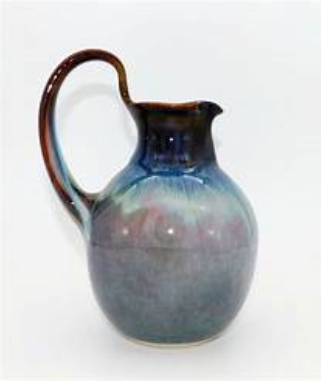 Fotografia przedstawia dzbanek szkliwiony. Dzbanek ma jeden duży uchwyt. Ma okrągły, pękaty kształt, ze zwężającą się szyjką. Duże, owalne ucho znajduje się po lewej stronie dzbana i sięga od środka, do brzegu szyjki. Od spodu dzban jest szary, następnie szarości przechodzą w róż, przy szyjce niebieski, u samej góry szyjki ciemnoniebieski.
