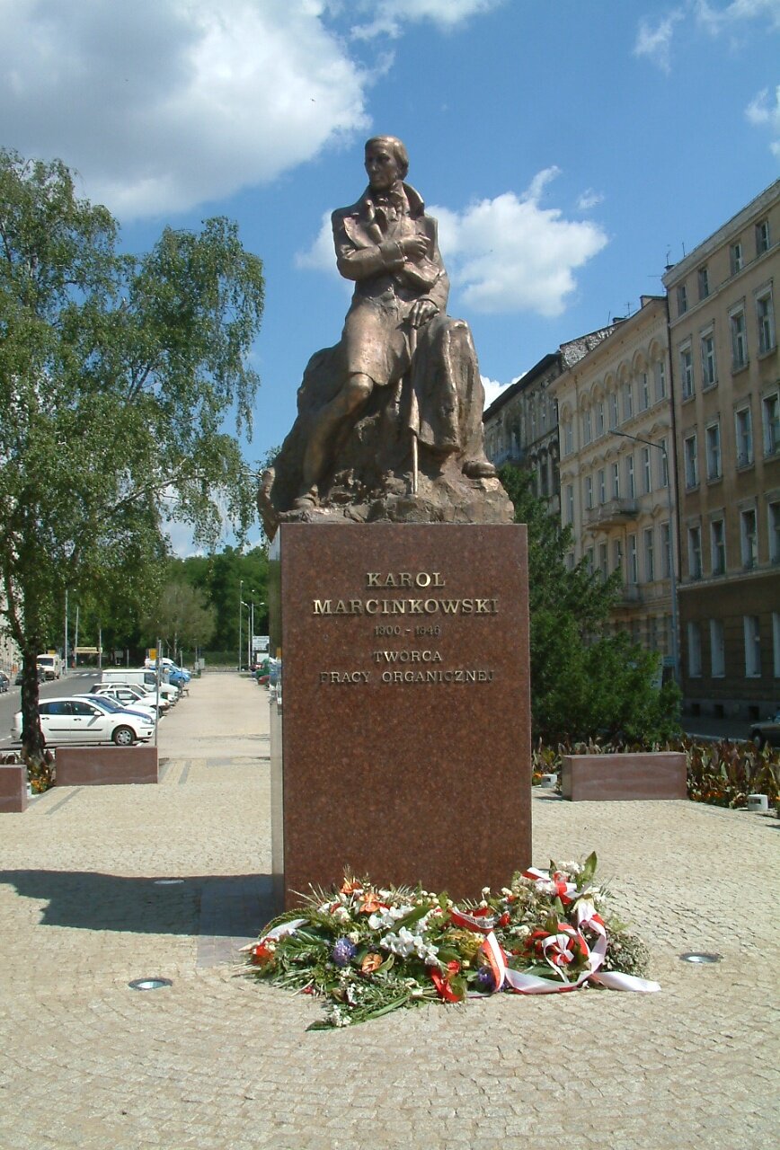 Kliknij, aby powiększyć Na zdjęciu pomnik, który przedstawia Karola Marcinkowskiego z laską w dłoni, w długim płaszczu z epoki. Mężczyzna przedstawiony w pozycji siedzącej. Napis na pomniku głosi: Karol Marcinkowski 1800 -1846 – twórca pracy organicznej. Odlana z brązu rzeźba Karola Marcinkowskiego umieszczona jest na cokole z czerwonego szwedzkiego granitu. Wysokość monumentu to 5 metrów. Cokół ma 2,2 metry wysokości, podstawa wymiary 1,8 x 1,8 metrów. Przed pomnikiem leżą świeże wiązanki kwiatów. Po prawej stronie zdjęcia kamienice, po lewej drzewo i parking. W tle drzewa, na niebie kilka chmur.