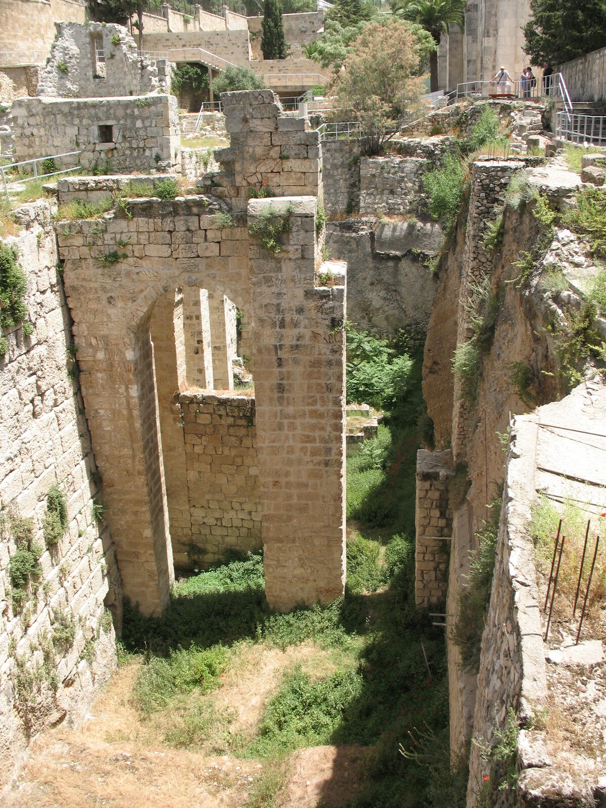 Kliknij, aby powiększyć. Na zdjęciu przedstawiono pozostałości zabudowań. Mur porośnięty jest trawą. Wokół murów widać współczesny chodnik, schody, barierki. Na chodniku, w oddali, w prawym górnym rogu zdjęcia, stoją ludzie.