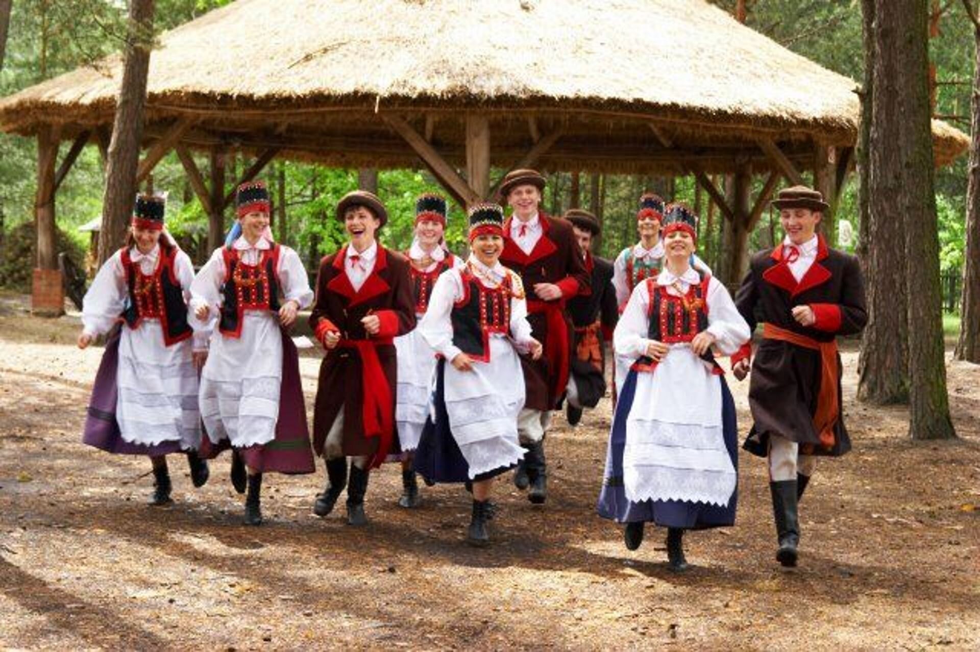 Fotografia przedstawia osoby z regionui Kurpiowskiego w tradycyjnych strojach. Uśmiechnięte kobiety w długich sukniach z białymi fartuszkami, na szyi mają czerwone korale. Mężczyźni tańczą obok kobiet w spodniach i długich płaszczach, a na głowie mają kapelusze.