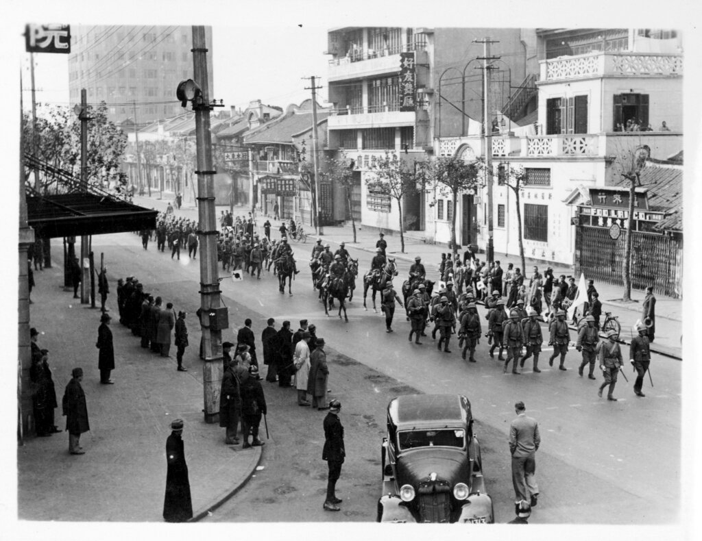 Ilustracja przedstawia ulicę, po której maszerują żołnierze. Wzdłuż ulicy stoją ludzie, oglądający defiladę wojsk.