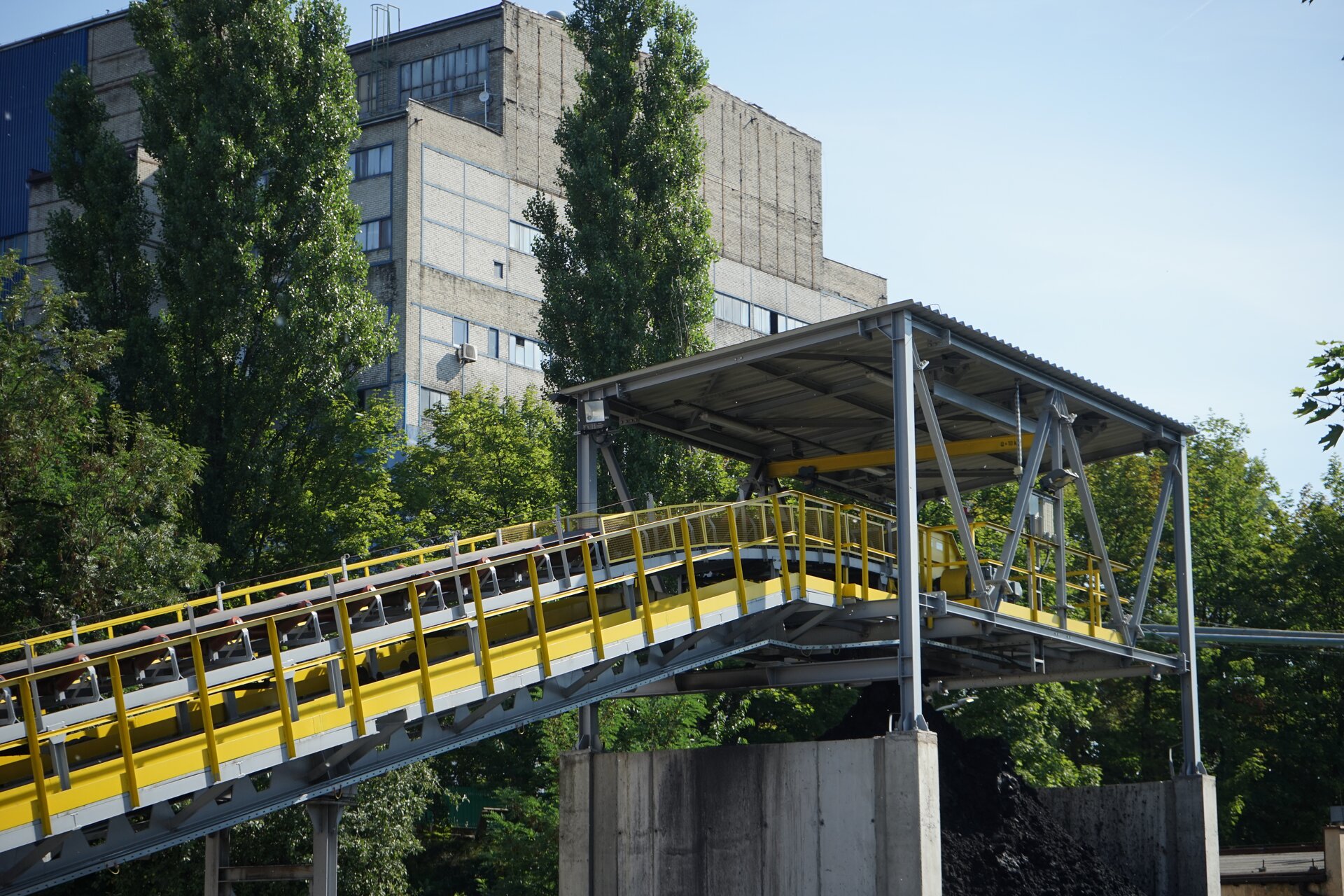 Na zdjęciu widoczny jest podwieszony przenośnik taśmowy. Przenośniki jest układami modułowym. Na stalowej prostopadłościennej ramię osadzone są lekko nachylonych rolki. Rolki mają kształt walca. Na rolkach znajduje się taśma. Boki przenośnika są zabezpieczone siatką oczkową. Przenośnik osadzony jest na stalowych filarach. Z prawej strony zdjęcia węgiel z przenośnika spada na hałdę.Poniżej zdjęcia widoczny jest przycisk multimedialny z nagraniem audio. Treść nagrania jest zgodna z tytułem zdjęcia.