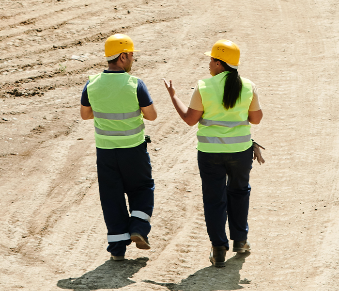 Zdjęcie drugie podpisane Określanie zasad obsługi codziennej i transportowej wskazanej maszyny. Zdjęcie przedstawia dwoje pracowników w kamizelkach odblaskowych i żółtych kaskach przechadzających się po placu budowy i rozmawiających ze sobą.