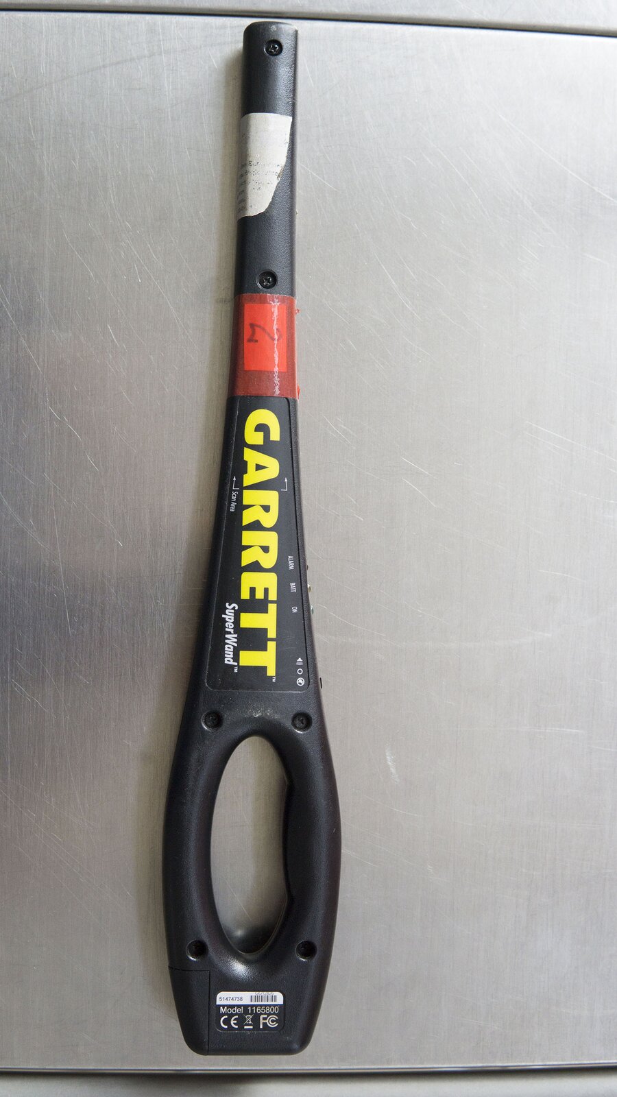 Zdjęcie przedstawia ręczny wykrywacz metalu do kontroli osób. Ma on kształt walca lekko rozszerzającego się w kierunku uchwytu. Częścią walcowatą wykonuje się kontrolę pasażera, przesuwając nią w powietrzu blisko ciała osoby sprawdzanej. Uchwyt detektora ma owalny kształt z owalnym wycięciem w środku tak, aby można było wygodnie przełożyć palce przez ten otwór, trzymając urządzenie.