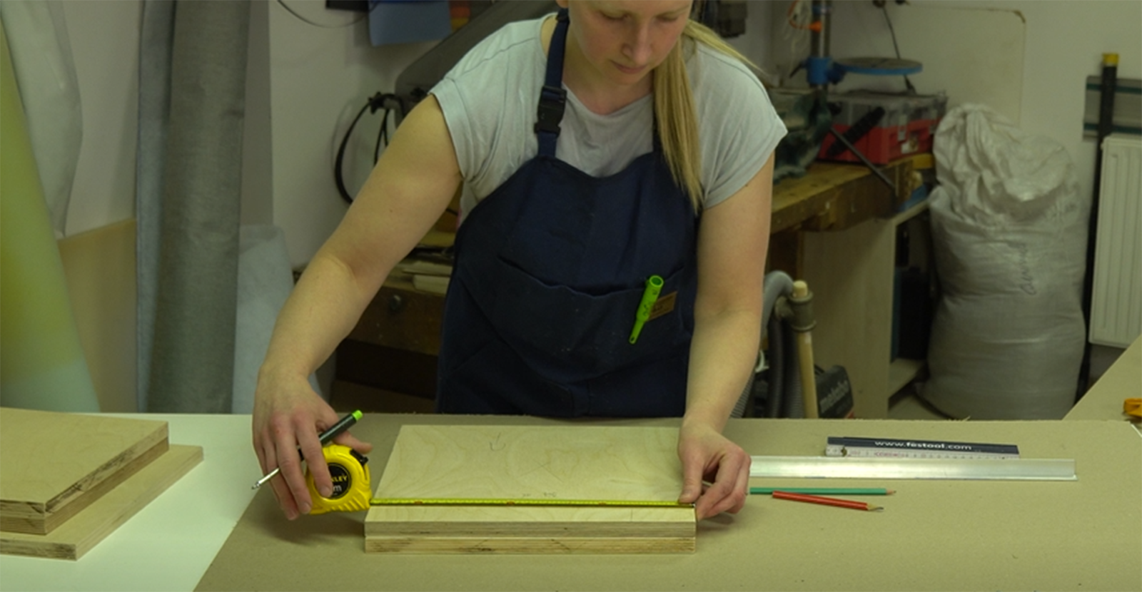 Zdjęcie przedstawia tapicera, który przygotowuje elementy drewnianej skrzynki, stanowiącej konstrukcję nośną pufa. Kobieta mierzy wycięty ze sklejki kwadratowy element.