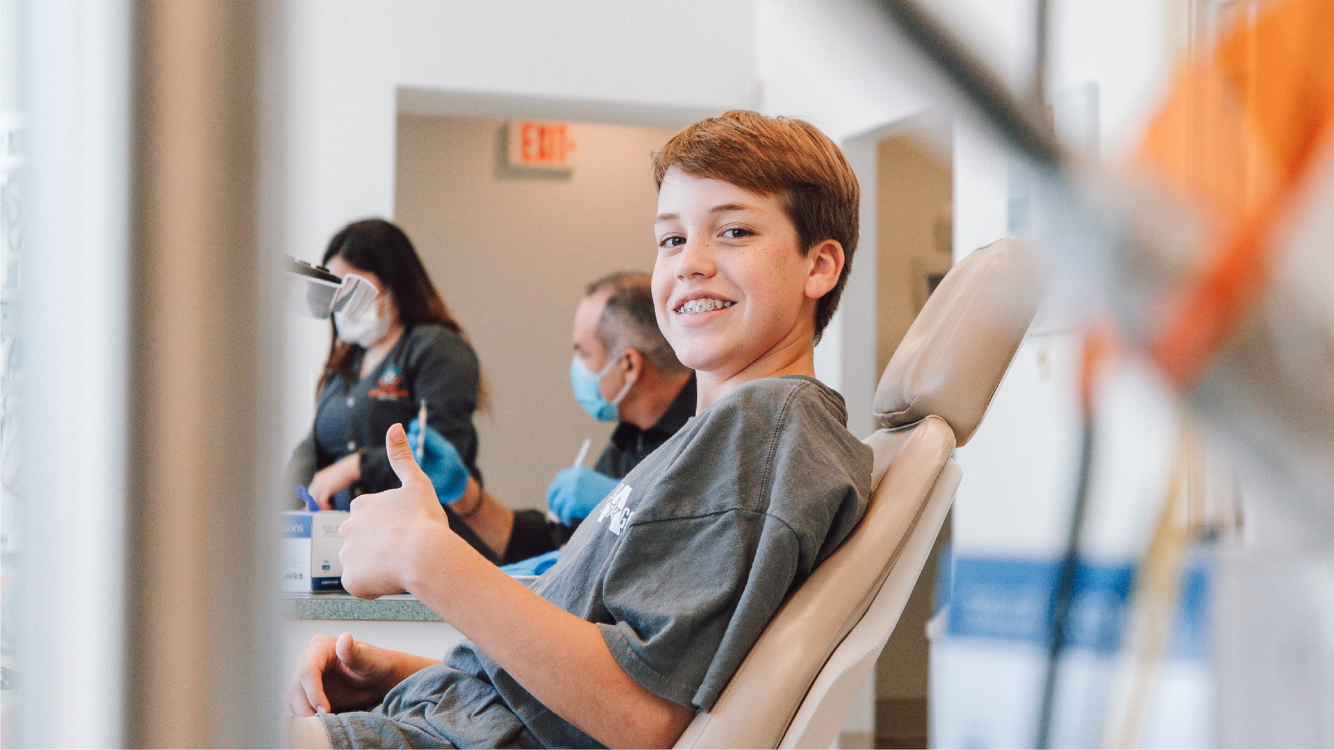 Zdjęcie przedstawia uśmiechniętego nastoletniego chłopca siedzącego na fotelu dentystycznym. Chłopiec unosi kciuk lewej dłoni w górę. W tle widać dwie osoby w maseczkach ochronnych.