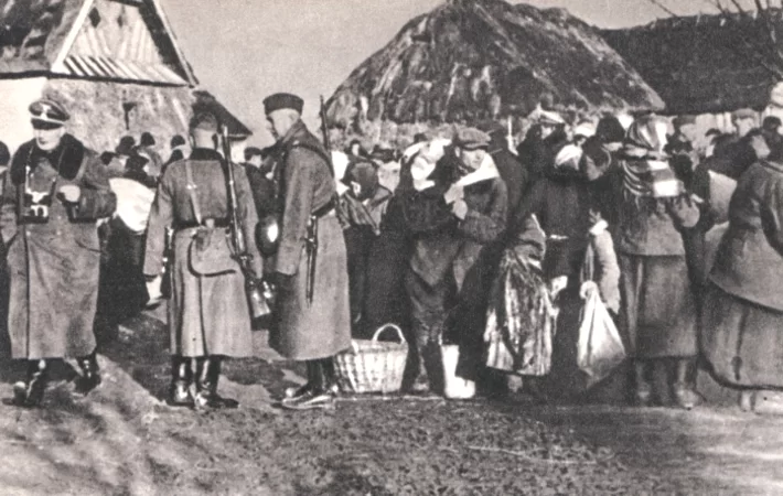 Kliknij, aby powiększyć Zdjęcie przedstawia dwóch niemieckich żołnierzy stojących z przewieszonymi przez ramię karabinami. Obok idzie niemiecki oficer w zawieszonym na szyi aparatem fotograficznym. Za nimi i po ich prawej stronie tłoczą się mężczyźni, kobiety i dzieci. Kobiety mają na głowach chusty. Niektórzy mają przy sobie tobołki, na ziemi stoi wiklinowy koszyk. Za ludźmi stoją dwie chaty, jedna kryta słomą.
