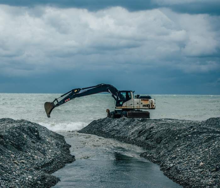 Zdjęcie podpisane Wykorzystywanie koparki podczas prac przekopowych do oceanu. Zdjęcie przedstawia koparkę znajdującą się na zwalisku ziemi i kamieni, przedzielonym wąskim przesmykiem, przez który przepływa woda. Za koparką widać ocean.