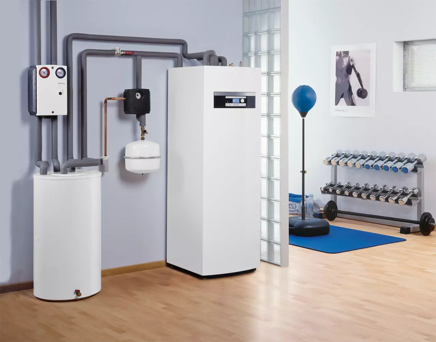 Zdjęcie przedstawia przykładowe miejsce zabudowy pompy ciepła wewnątrz budynku. Po lewej stronie na podłodze stoi zbiornik na wodę, który ma kształt walca. Nad nim znajduje się urządzenie w obudowie w kształcie prostopadłościanu. Zabudowano w nim grupę pomp obiegowych z zaworami. Na prawo zawieszony jest zbiornik wyrównawczy mający postać niewielkiej beczki. Po prawej stronie zaś widoczne jest urządzenie jednostki wewnętrznej pompy. Jego obudowa ma kształt dużego prostopadłościanu z niewielkim panelem sterującym oraz wyświetlaczem w górnej części. Wszystkie elementy instalacji są połączone systemem rur zabezpieczonych szarą otuliną.