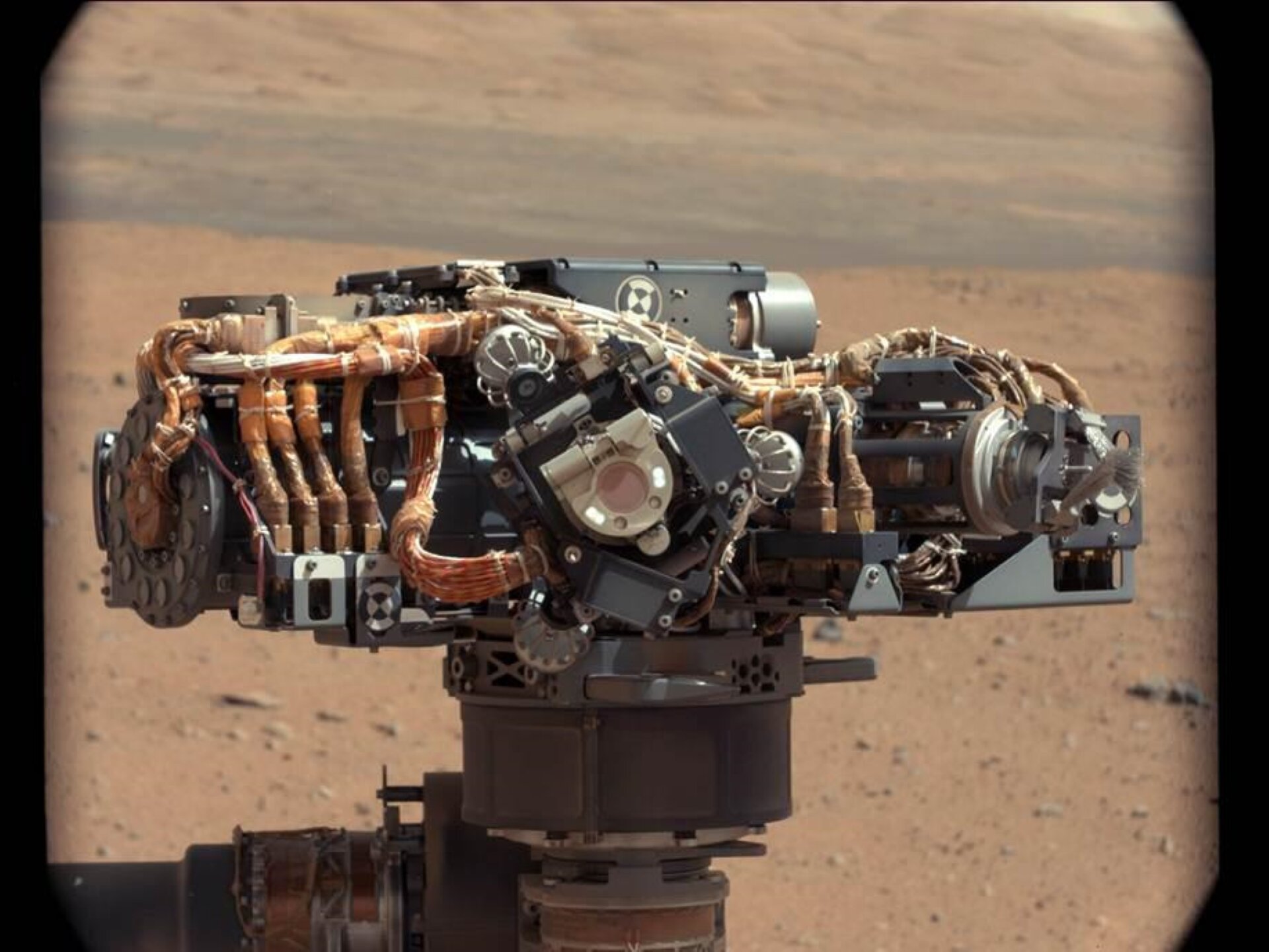 Rys. 6. górny. Ilustracja przedstawia zdjęcie łazika Curiosity badającego powierzchnię Marsa od dwa tysiące dwunastego roku. Na zdjęciu pokazano niewielki pojazd kołowy przemierzający piaszczystą powierzchnię Marsa. Nad podstawą pojazdu widoczna jest pionowa kolumna, na zwieńczeniu której znajdują się liczne urządzenia pomiarowe. Systemy pomiarowe widoczne są w postaci wiązek przewodów, chwytaków, kamer i licznych innych urządzeń. 