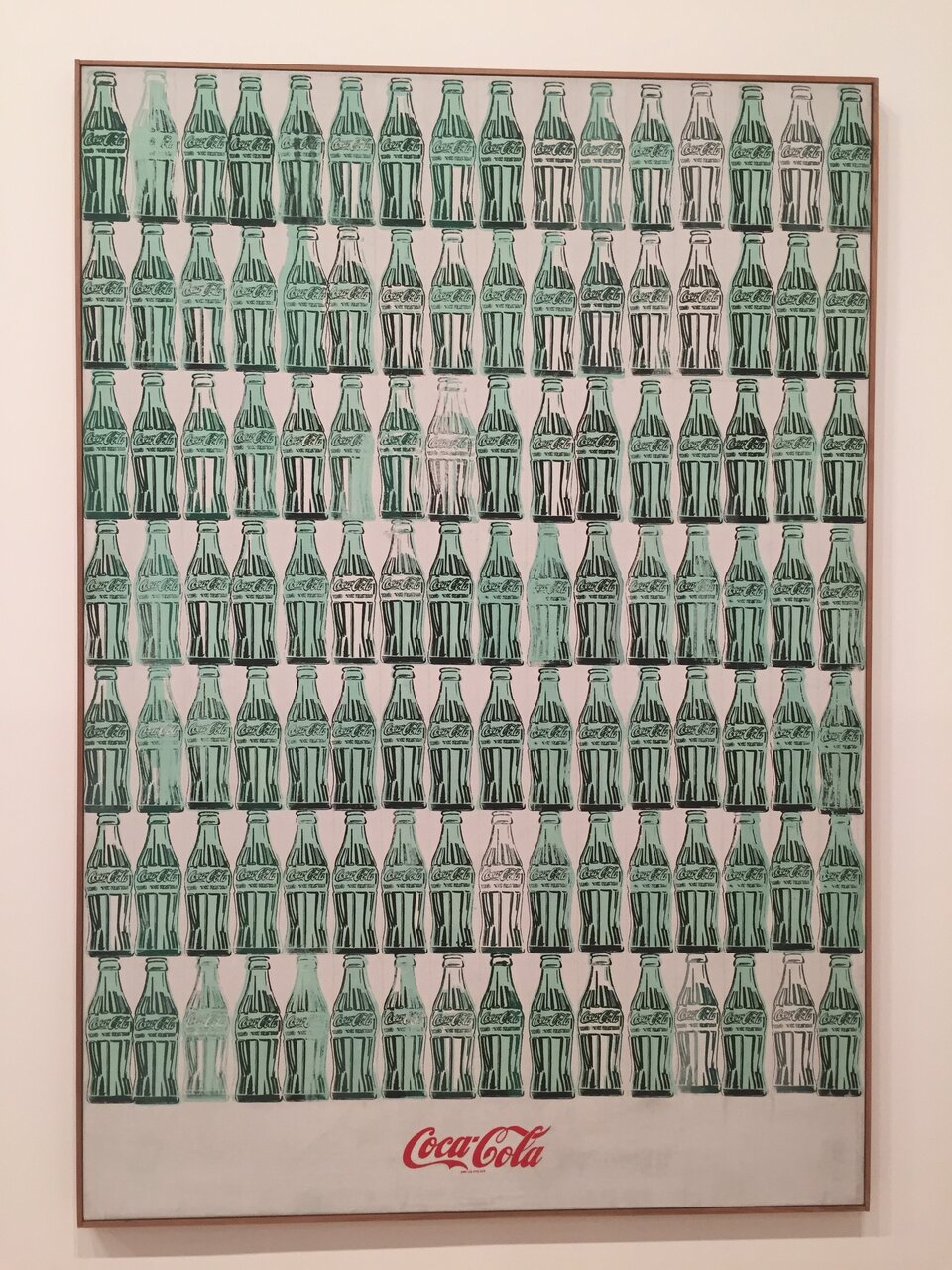 Ilustracja przedstawia dzieło Andy'ego Warhola „Butelki Coca‑Coli” o kształcie pionowego prostokąta. Ukazuje ułożone w pionie szesnaście zielonych butelek ustawionych w rzędzie, powielonych w pionie siedem razy. Na dole, pod butelkami, znajduje się czerwone logo z napisem Coca Cola. Tło grafiki jest białe.